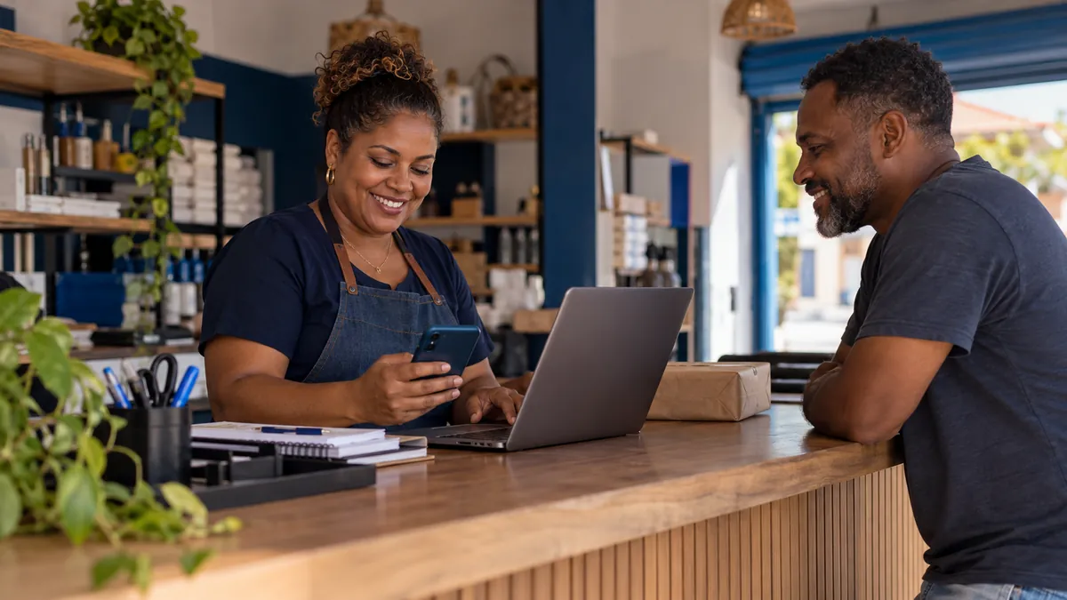 Dona de pequeno negócio em Conceição do Jacuípe organiza pedidos no notebook e no celular no balcão, enquanto conversa com um cliente conhecido.