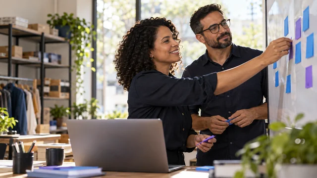 Dupla comercial brasileira organiza oportunidades em quadro físico com cartões azuis e violetas sem palavras, notebook no primeiro plano, pequena empresa organizada.